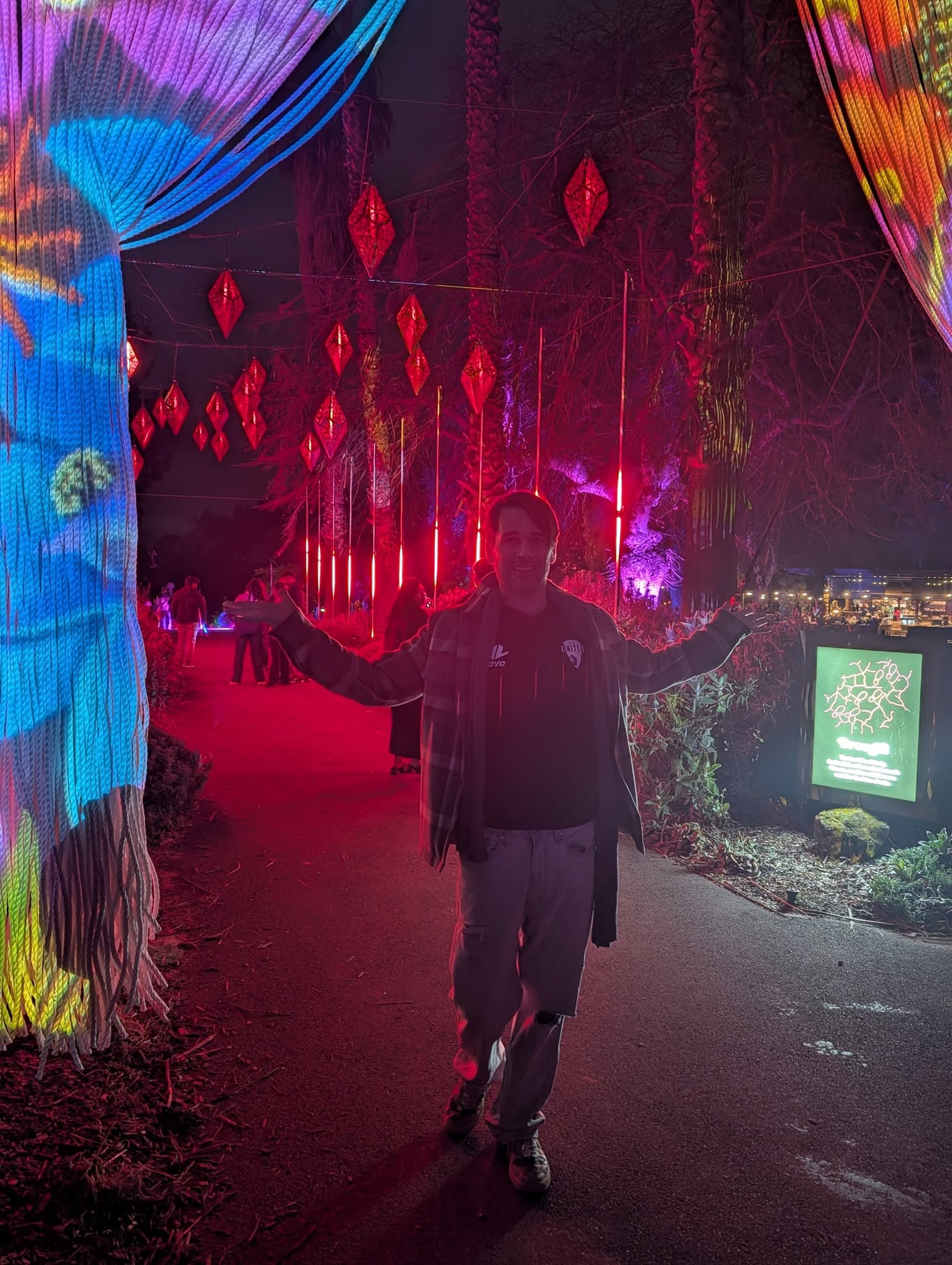 David Greenwood OAM standing with arms outstretched beneath red hanging lanterns and illuminated woven fabric at the Geelong Botanic Gardens light experience