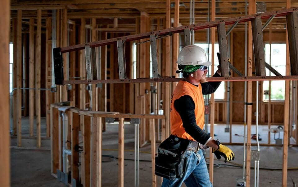 A builder carrying a ladder through the timber framing of a house under construction
