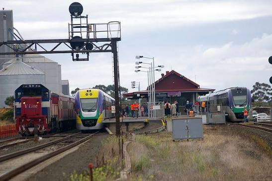 Three V/Line trains at Lara Station with passengers on the platform and grain silos behind