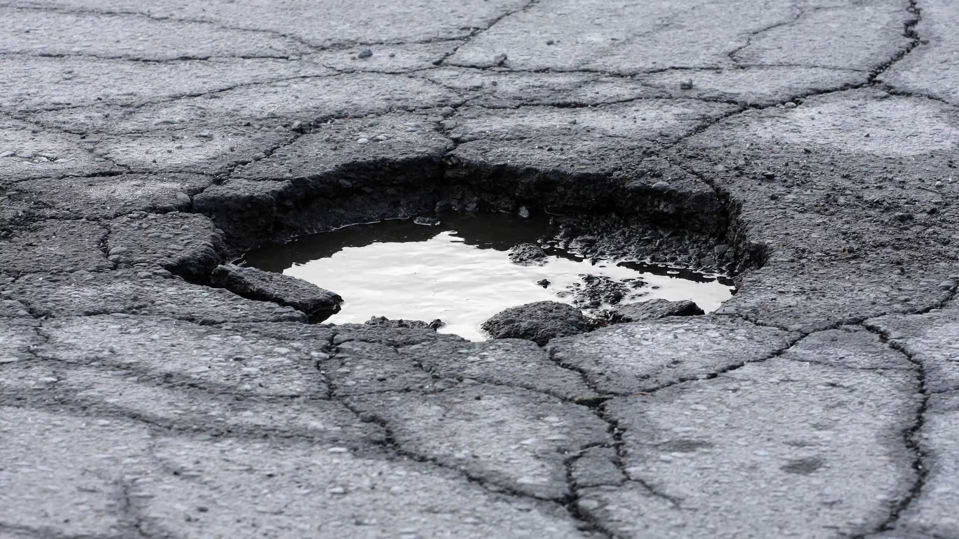 A large water-filled pothole in cracked asphalt on a suburban road