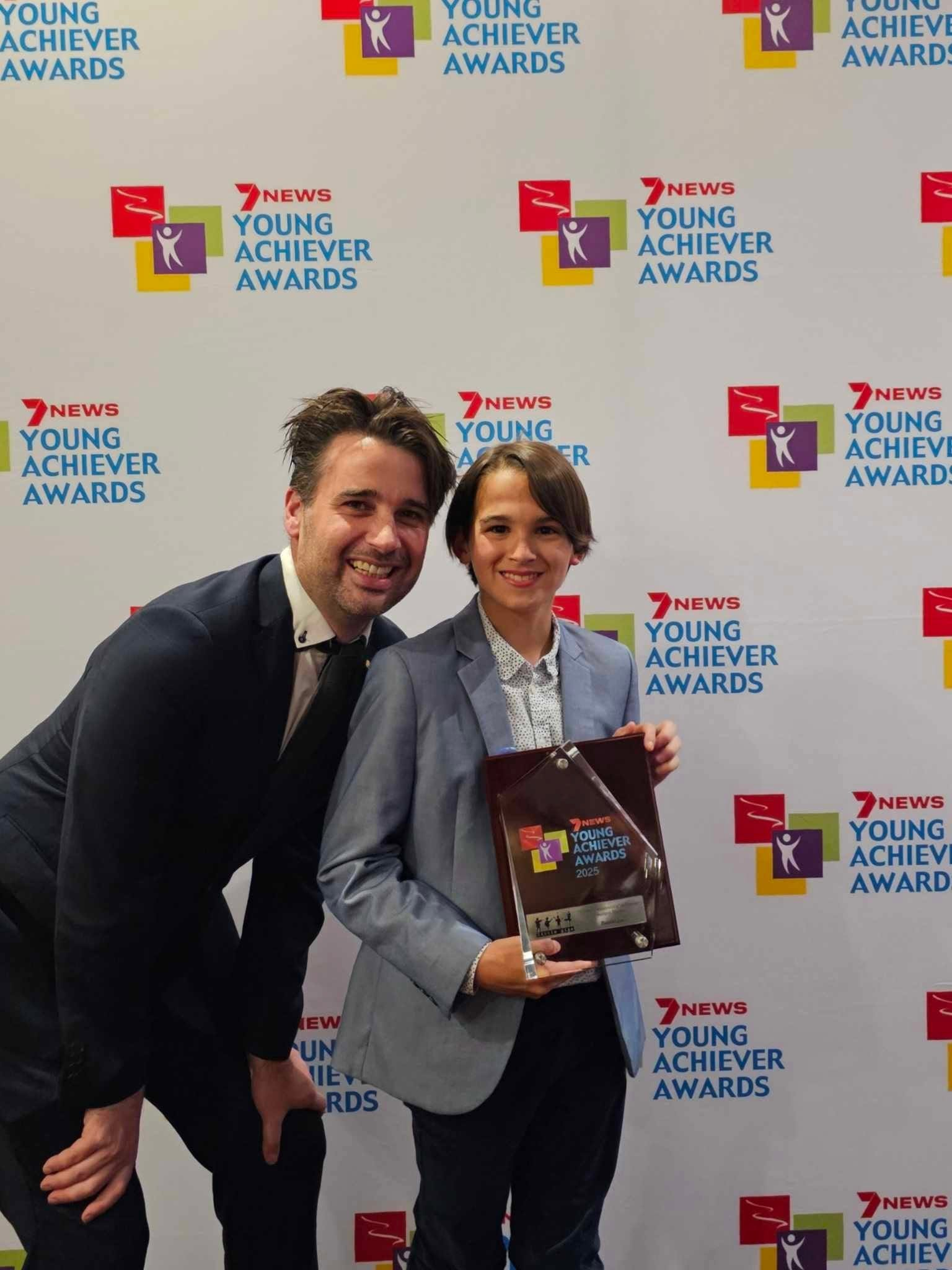 David Greenwood OAM standing with Daniel, who is holding his 2025 7NEWS Young Achiever Award, in front of the awards backdrop